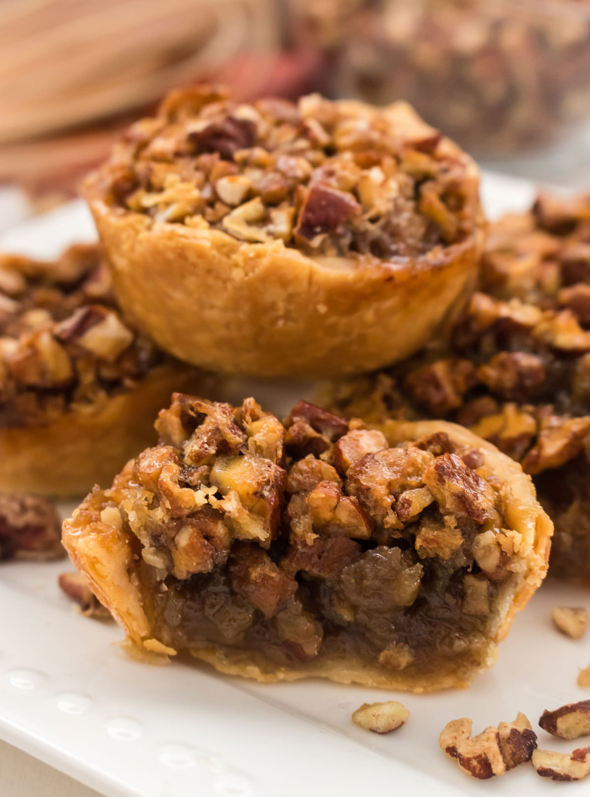 Closeup on five Mini Pecan Pies sitting on a white plate in front of a glass bowl filled with chopped pecans.