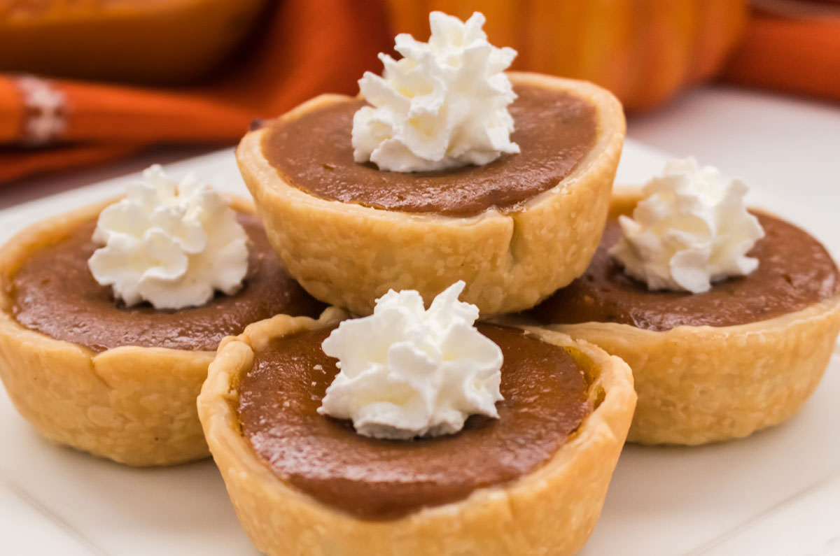 Closeup on a stack of five Mini Pumpkin Pies, topped with dollops of whipped cream, sitting on a white plate.