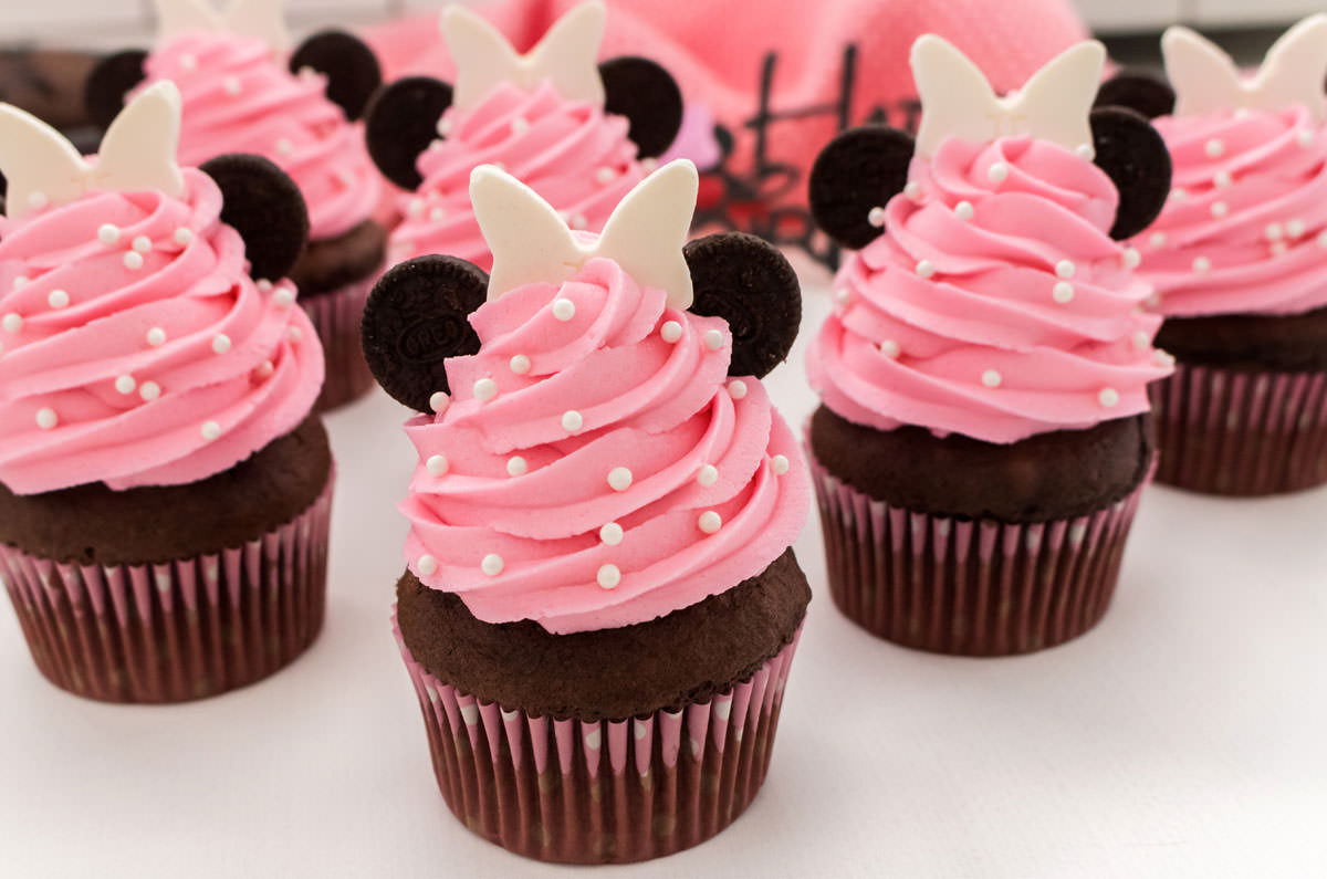 Closeup on six Minnie Mouse Cupcakes sitting on a white surface in front of a pink towel and a Happy Birthday sign.