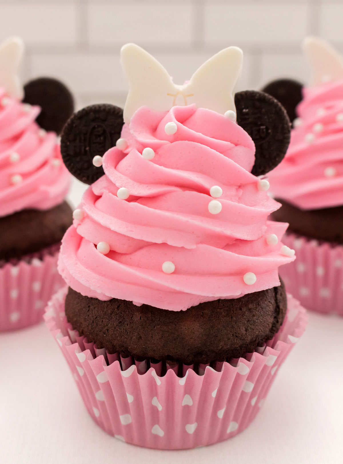 Closeup of a pink Minnie Mouse Cupcake with pink buttercream frosting and Oreo cookie ears sitting on a white table in front of other cupcakes.