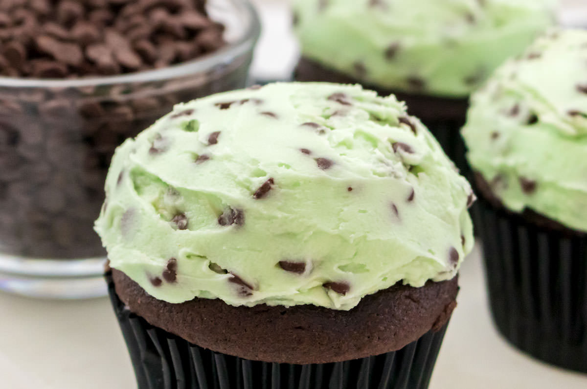 Closeup on cupcakes topped with Mint Chocolate Chip Buttercream Frosting sitting on a white surface next to a ramekin filled with Mini Chocolate Chips.