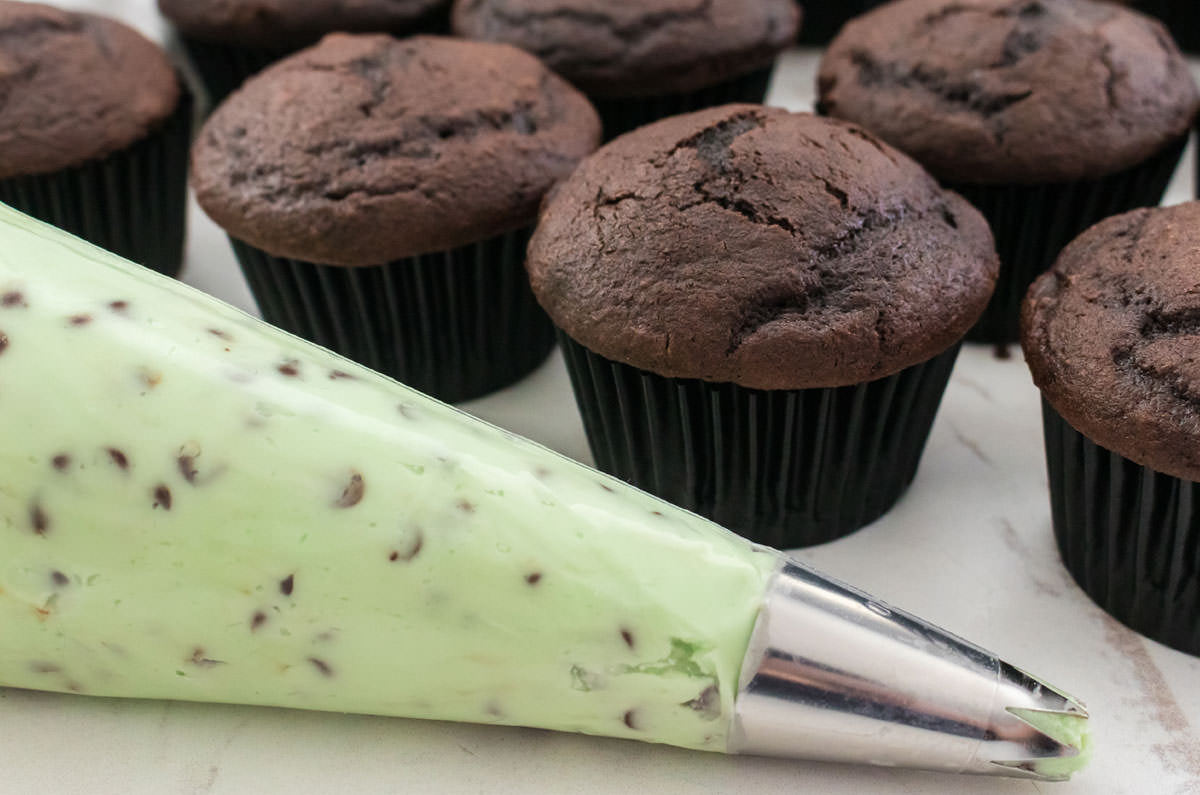 Closeup on a decorating bag filled with Mint Chip Frosting sitting in front of a batch of unfrosted chocolate cupcakes.