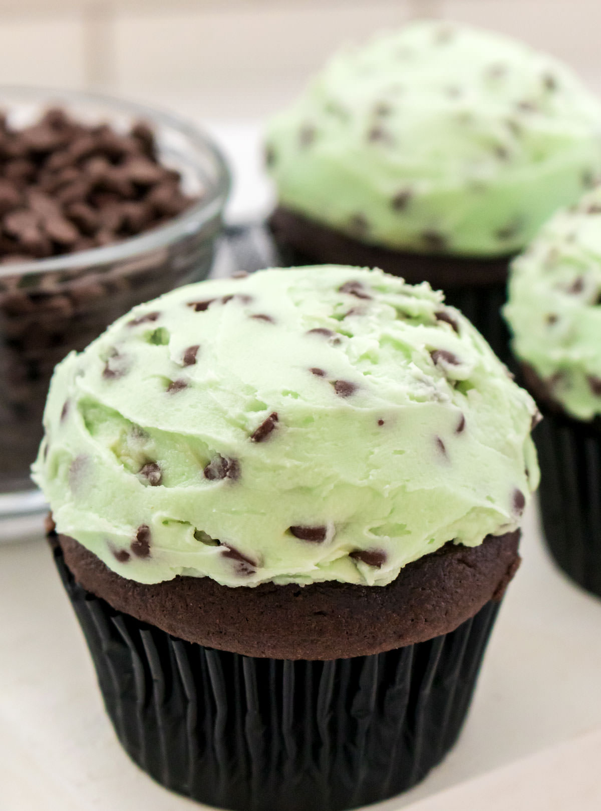 Closeup on three chocolate cupcakes frosted with Mint Chocolate Chip frosting sitting on a white table next to a bowl filled with chocolate chips.