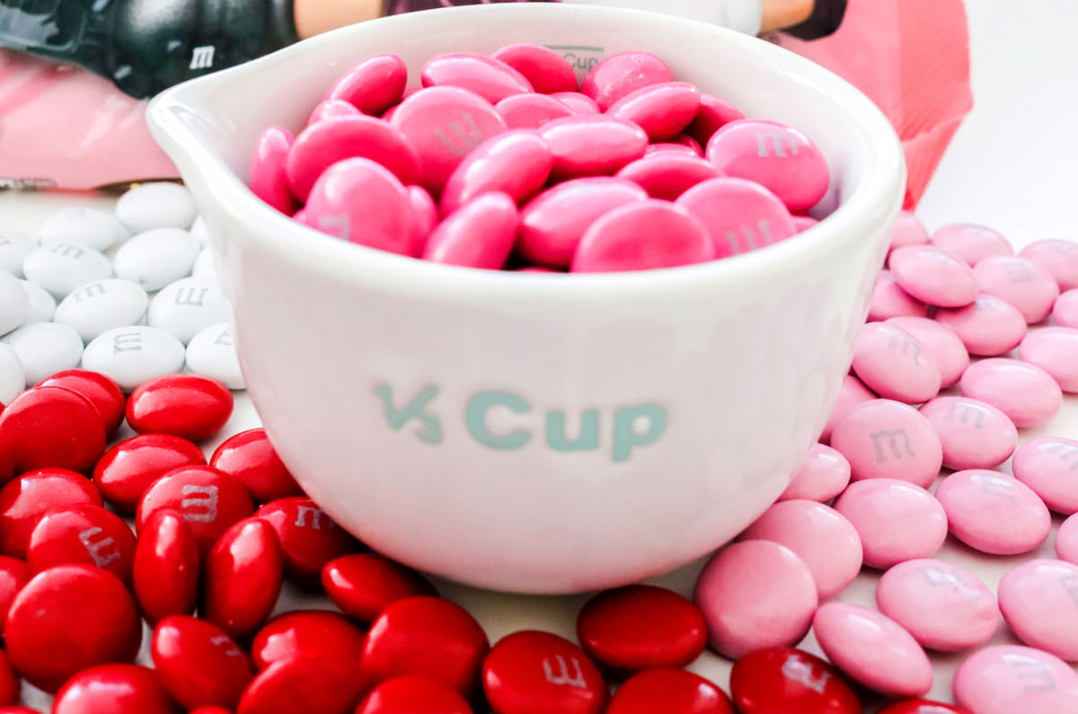 White measuring cup holding dark pink M&M's surrounded by red, light pink and white M&M's.
