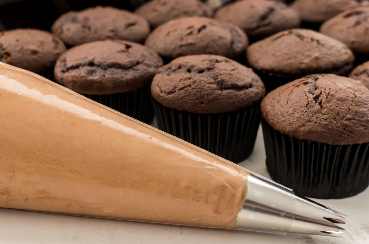 Closeup on a decorating bag filled with Mocha Frosting sitting in front of a batch of unfrosted chocolate cupcakes.