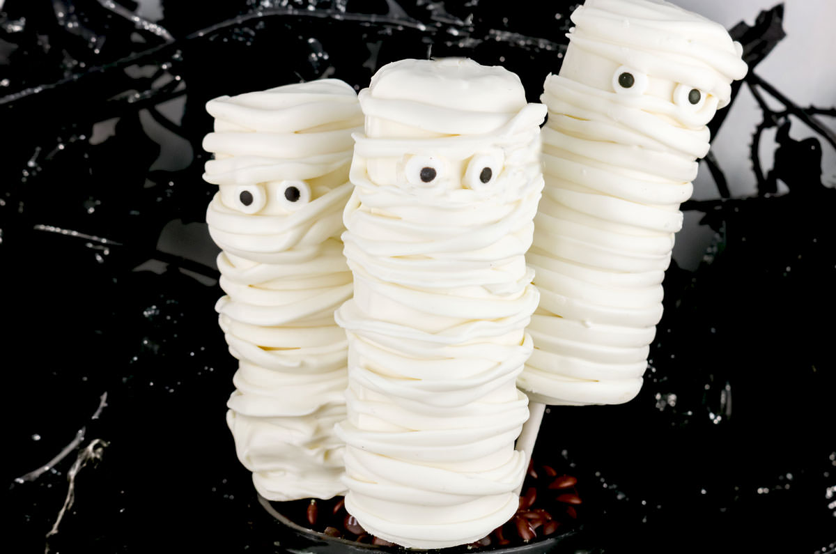 Closeup on three Mummy Marshmallow Pops propped up in front of a spooky black background.