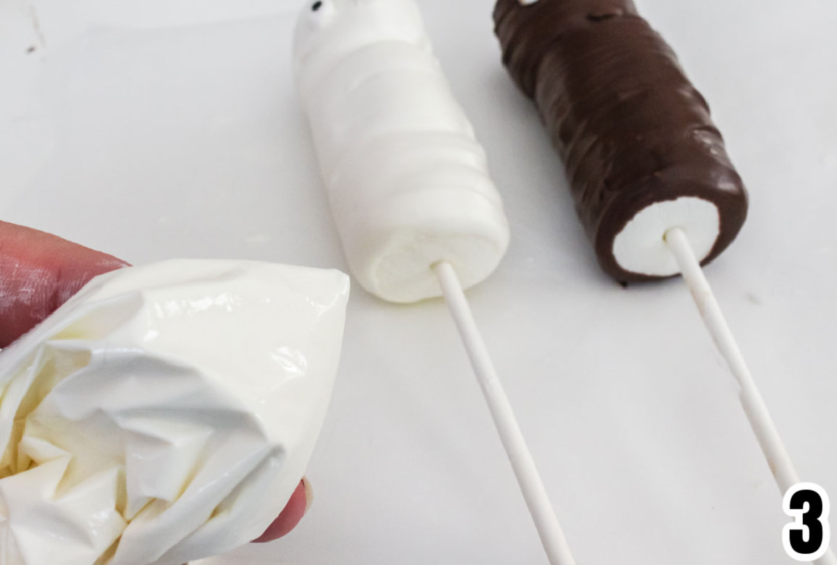 Two Mummy Marshmallow Pops laying on a white table with a plastic bag filled with melted White Candy Melts.