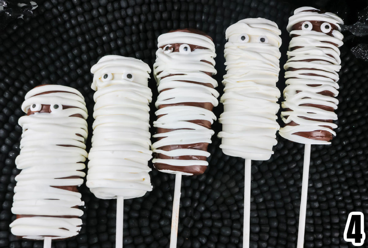 Five Mummy Marshmallow Pops laying on a black placemat.