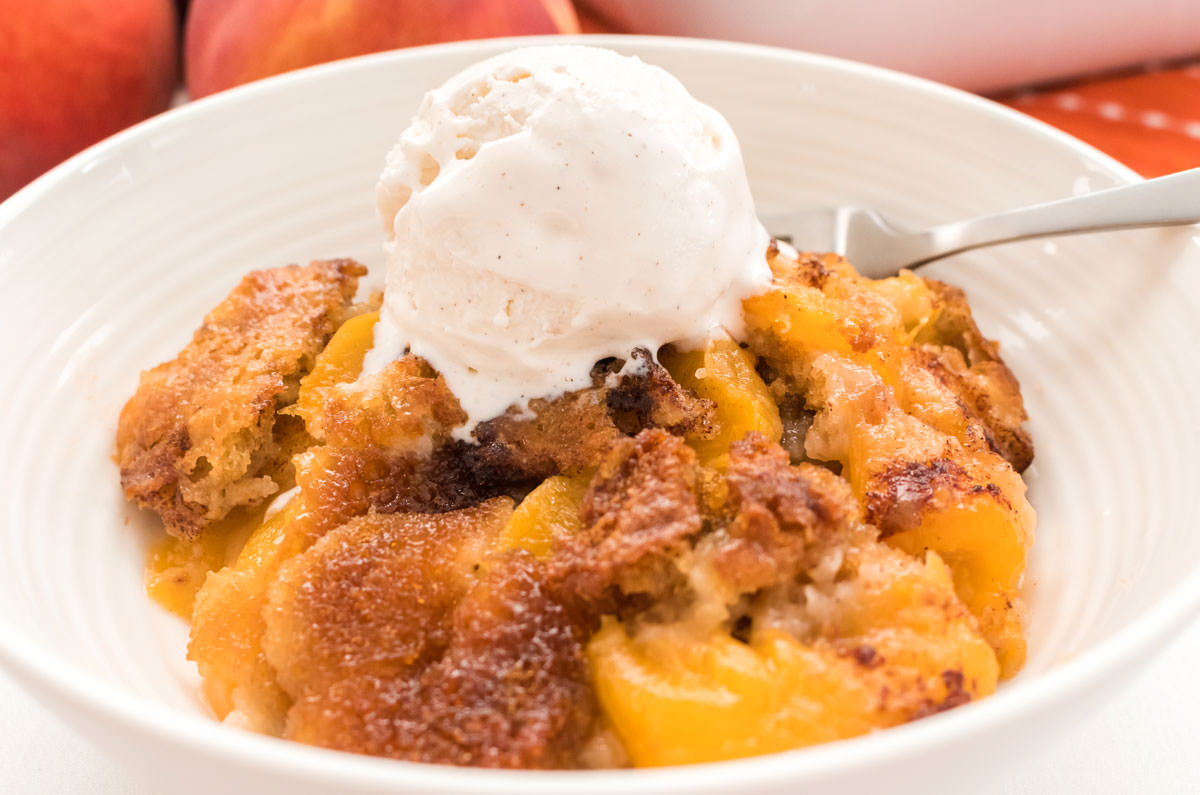 Closeup on a white bowl filled with Old Fashioned Peach Cobbler and a scoop of vanilla ice cream sitting in front of a stack of fresh peaches.