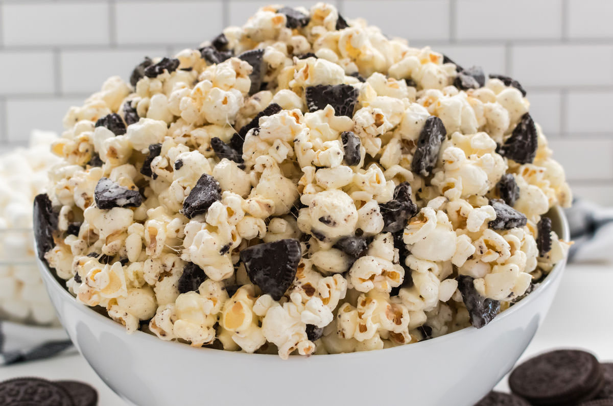 Closeup on a white serving bowl filled with Oreo Popcorn sitting on a white table surrounded by Oreo Cookies.