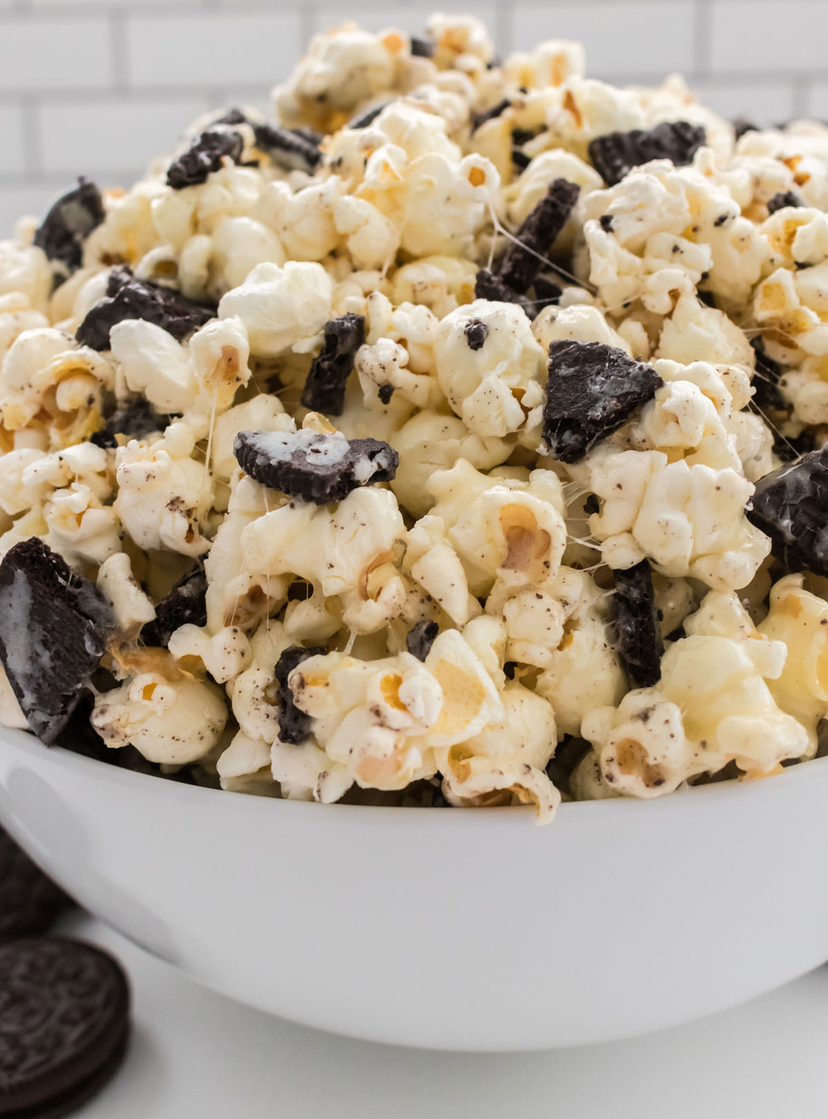 Closeup on a white serving bowl filled with Oreo Popcorn sitting on a white table surrounded by Oreo Cookies.