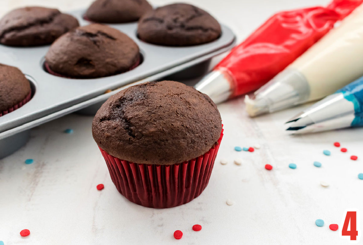 Chocolate cupcake in a red cupcake liner sitting on a white table alongside  three decorating bags filled with frosting and a cupcake tin filled with cupcakes.