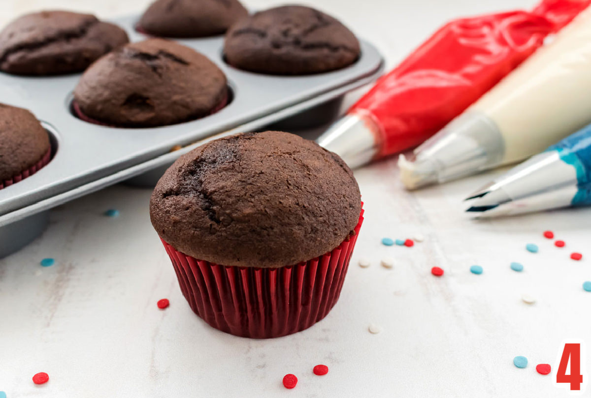 Chocolate cupcake in a red cupcake liner sitting on a white table alongside  three decorating bags filled with frosting and a cupcake tin filled with cupcakes.