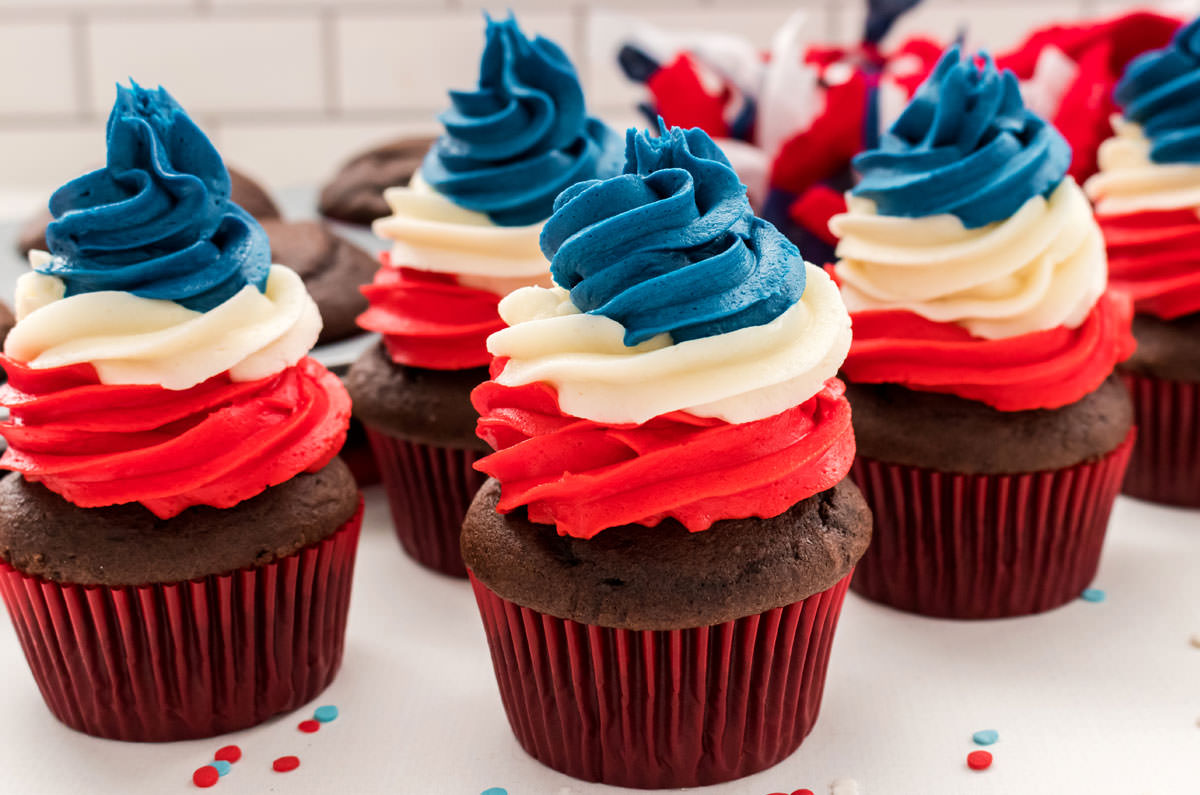 Closeup on five Patriotic Swirl Cupcakes sitting on a white table in front of a cupcake tin filled with chocolate cupcakes.