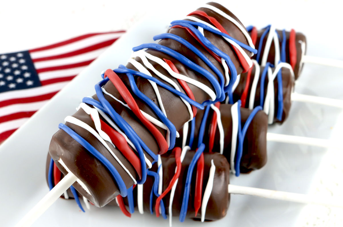 Closeup on a Patriotic Marshmallow Pop being held up in front of a serving platter full of more marshmallow pops and a mini American Flag.