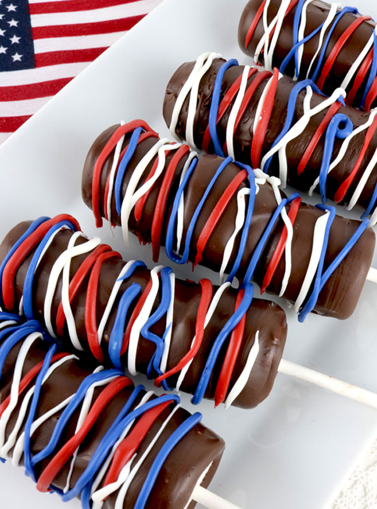 Closeup on five Patriotic Marshmallow Pops laying on a white serving platter next to an American Flag.