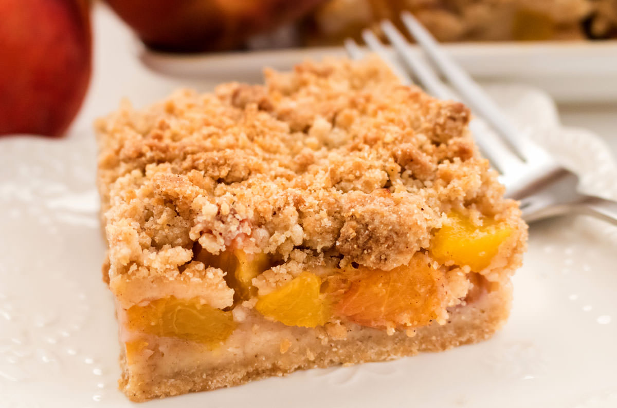 Closeup on a single Peach Crumble Bar on a white plate with a fork with peaches and more cookie bars in the background.