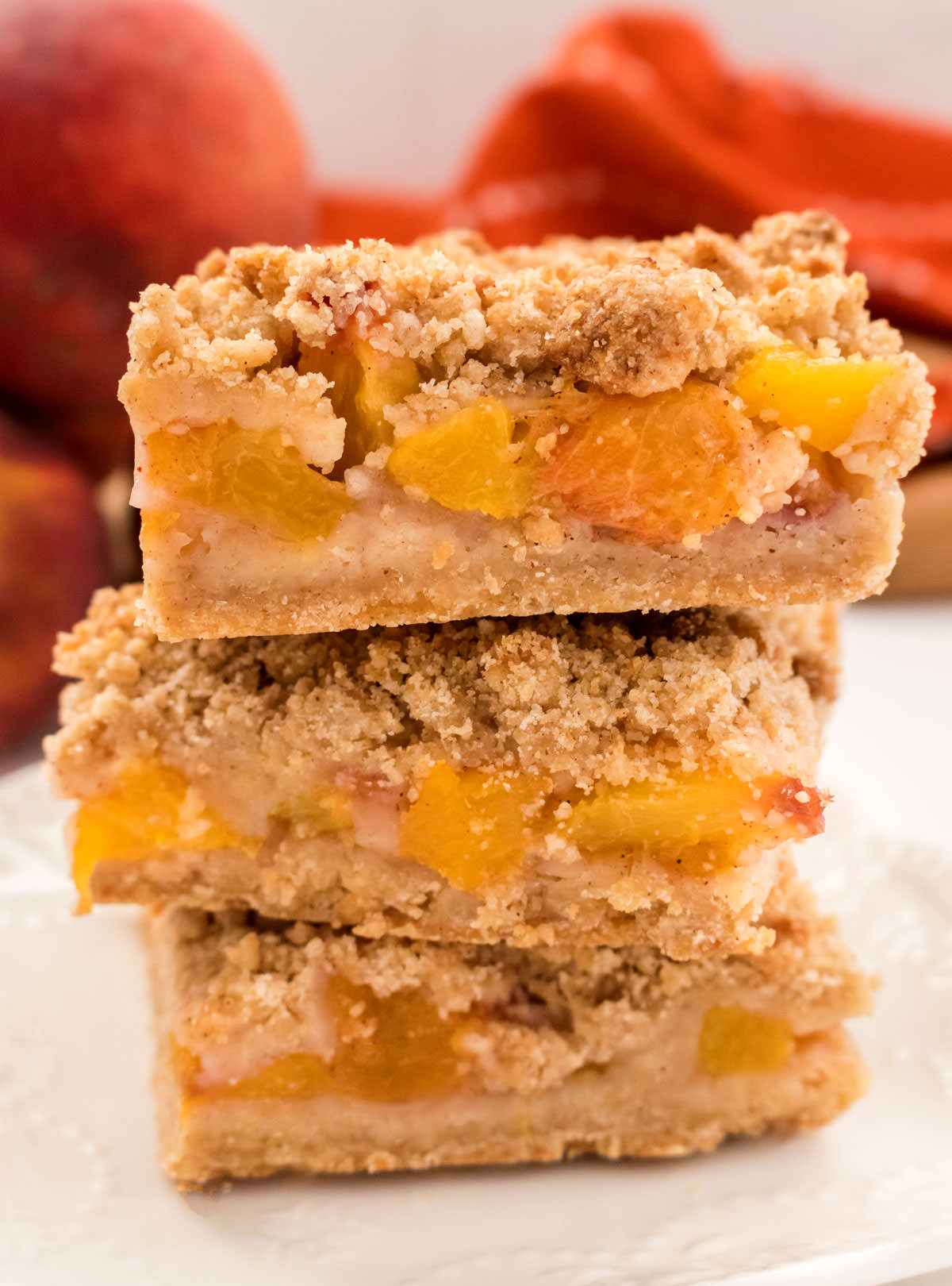 Closeup on a stack of Peach Crumble Bars sitting on a white plate in front of a orange towel and three fresh peaches.