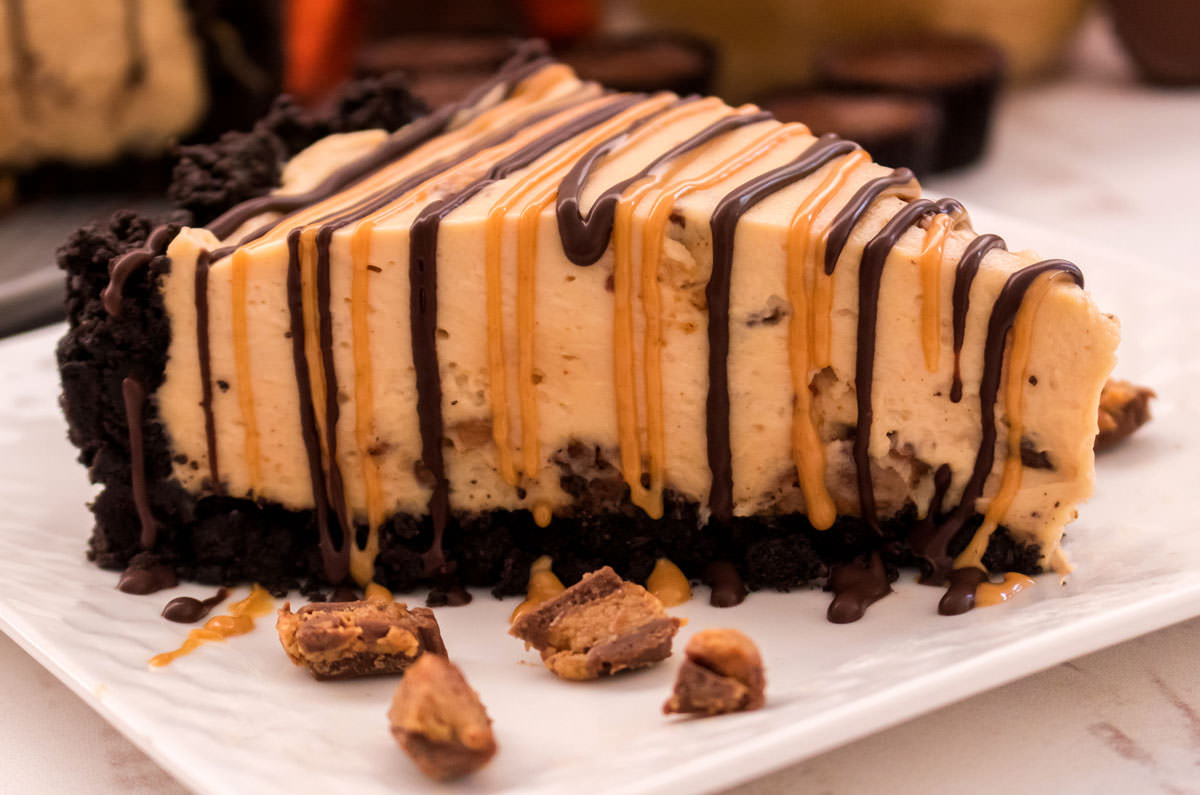 Closeup on a slice of No Bake Peanut Butter Cheesecake sitting on a white plate surrounded by pieces of Reese's Peanut Butter Cups.