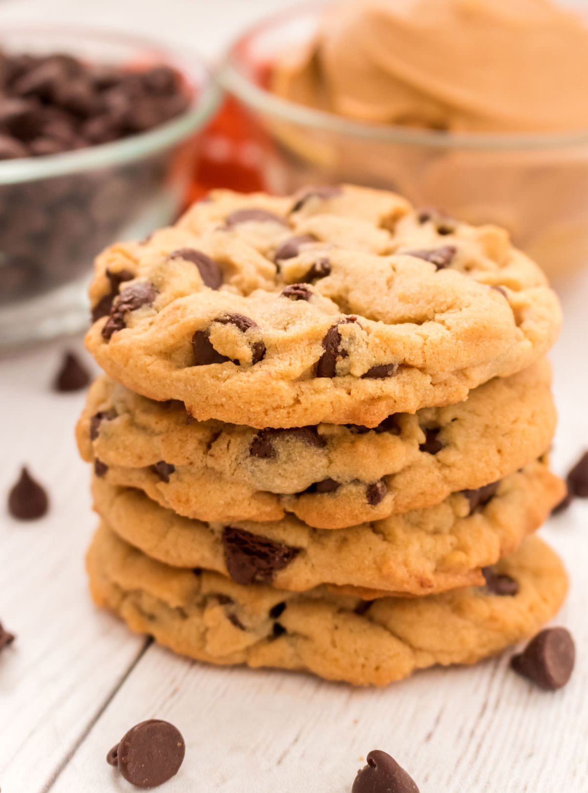 Closeup on a stack of Peanut Butter Chocolate Chip Cookies sitting on a white table in front of glass bowls filled with peanut butter and chocolate chips.