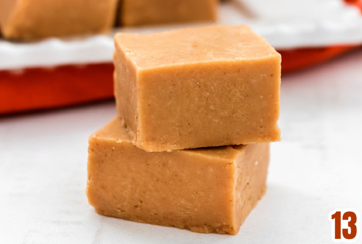Closeup on a stack of two pieces of Peanut Butter Fudge sitting on a white surface in front of a white platter filled with pieces of fudge.
