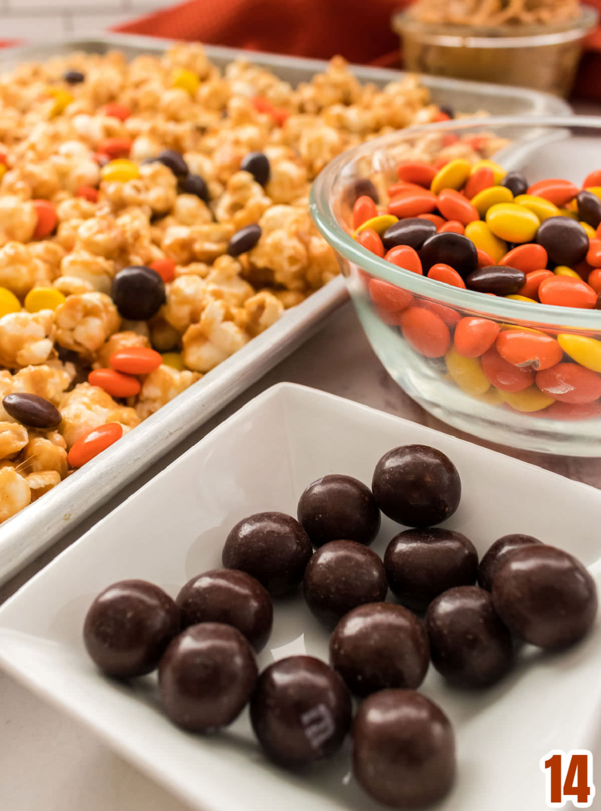Peanut Butter Popcorn laying flat in a cookie sheet sitting next to a ramekin filled with Brown Pretzel M&M's and a glass bowl filled with Reese's Pieces candy.