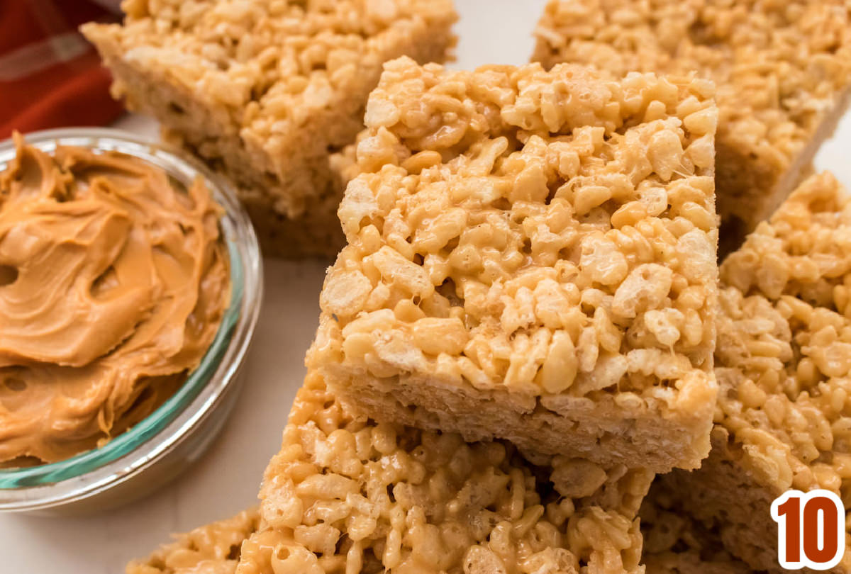 Overhead shot of a dozen Peanut Butter Rice Krispie Treats spread out on a white surface next to a bowl of peanut butter.