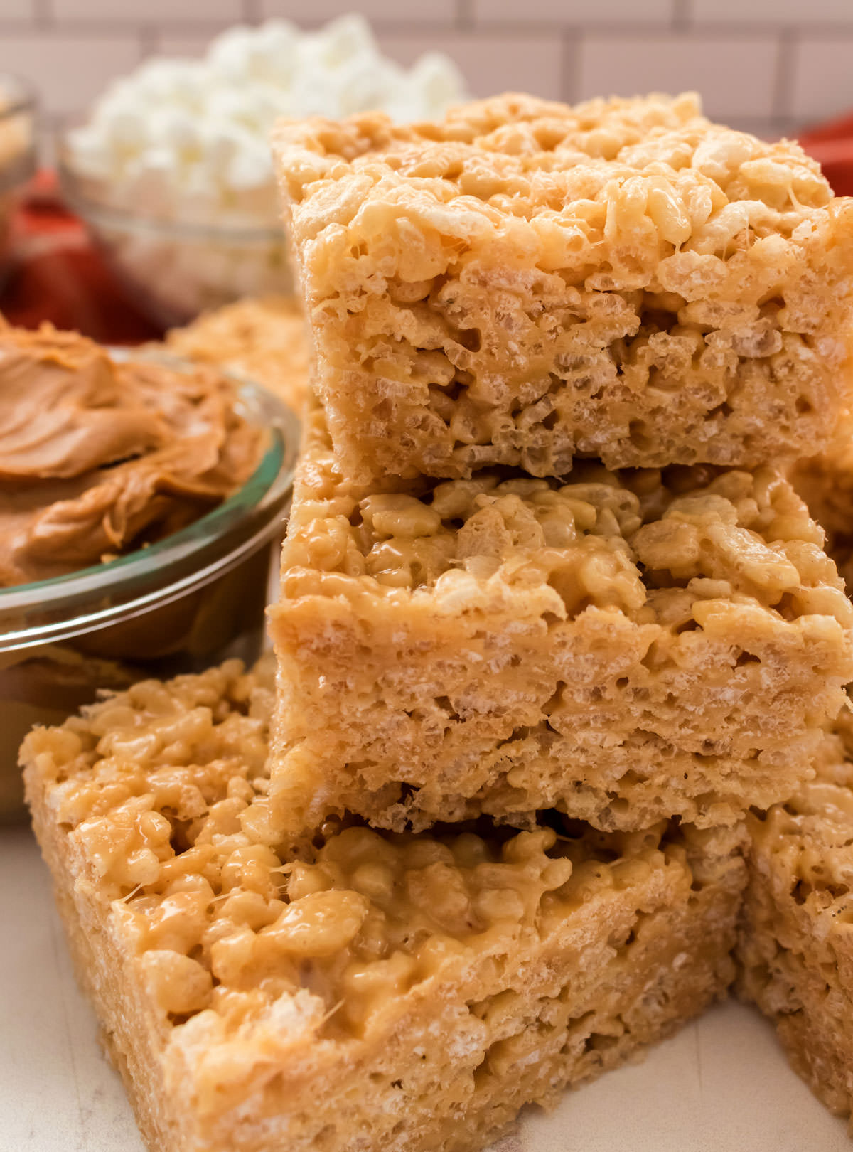 Closeup on a stack of three Peanut Butter Rice Krispie Treats sitting on a white surface next to a glass bowl of peanut butter.