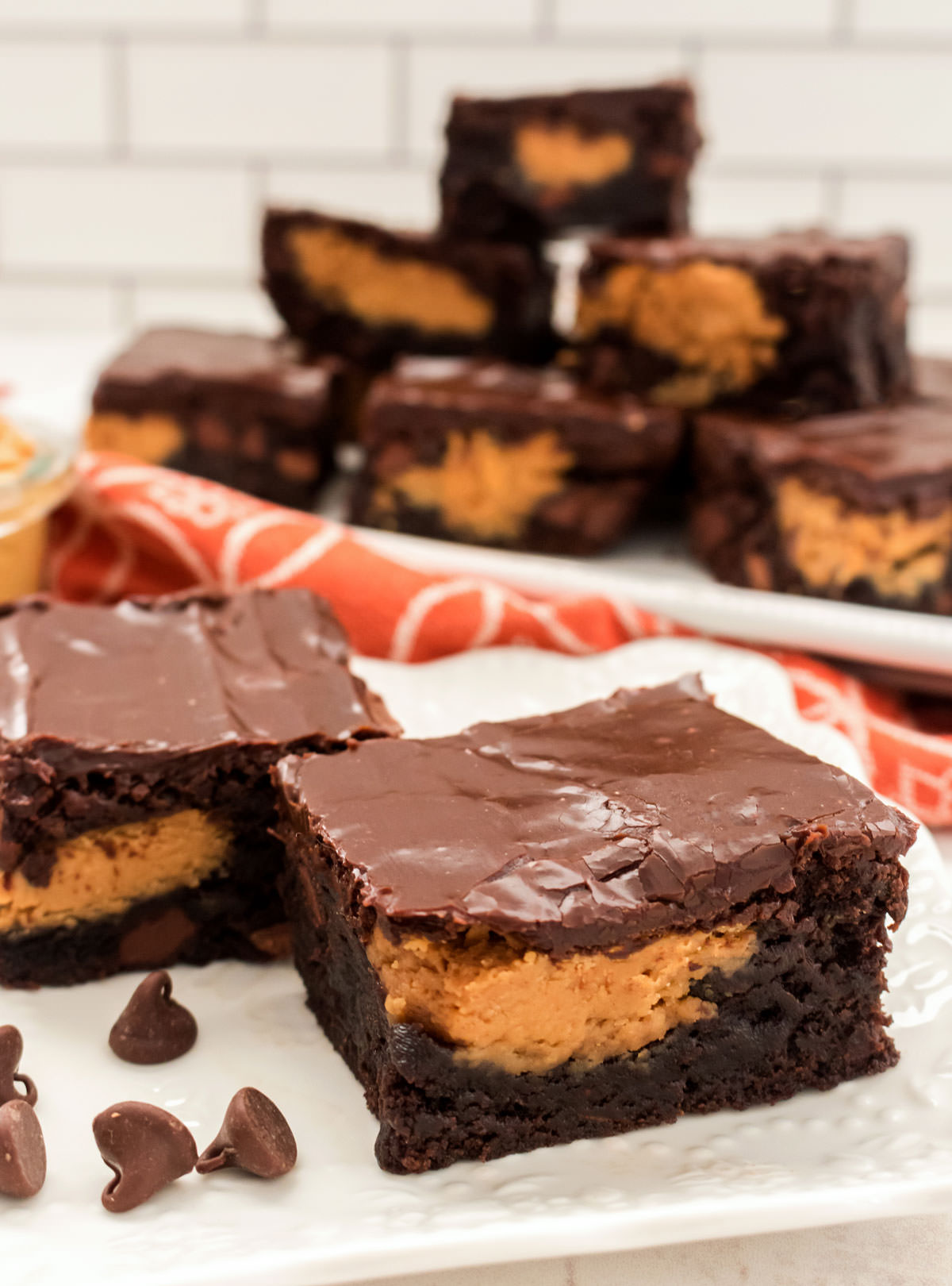 Two brownies sitting on a white plate in front of a platter full of Peanut Butter Swirl Brownies.