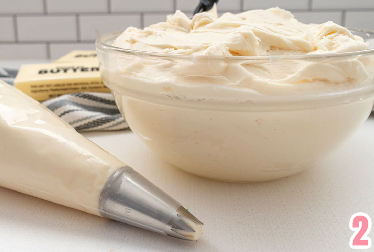 Closeup on a clear glass bowl filled with homemade buttercream frosting sitting next to a piping bag filled with frosting.