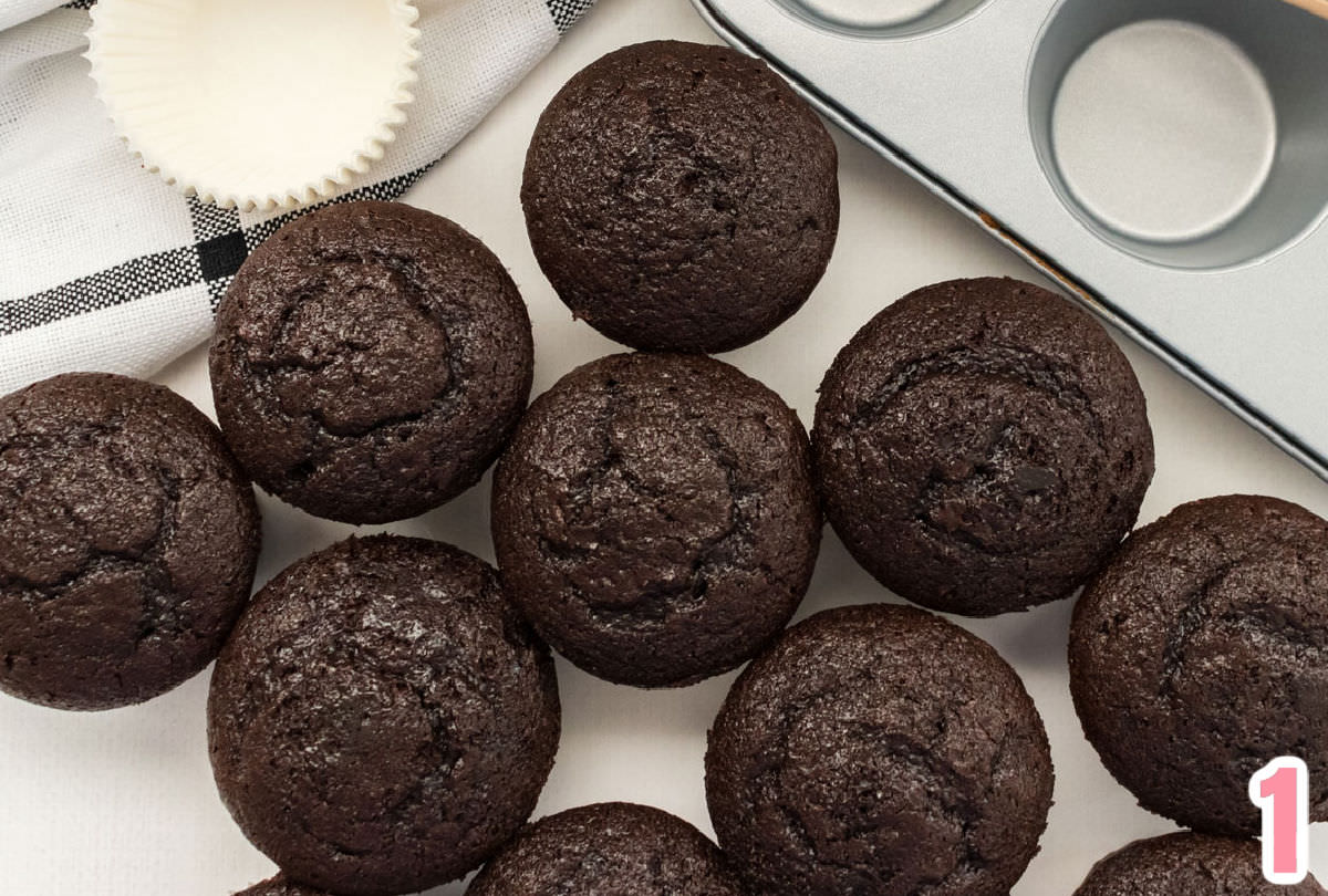 12 chocolate cupcakes sitting on a white table in front of a cupcake tin and a black and white towel.