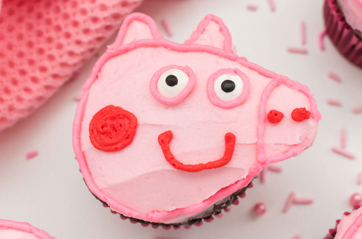 Closeup on a Peppa Pig Cupcake sitting on a white surface surrounded by pink sprinkles and a pink towel.