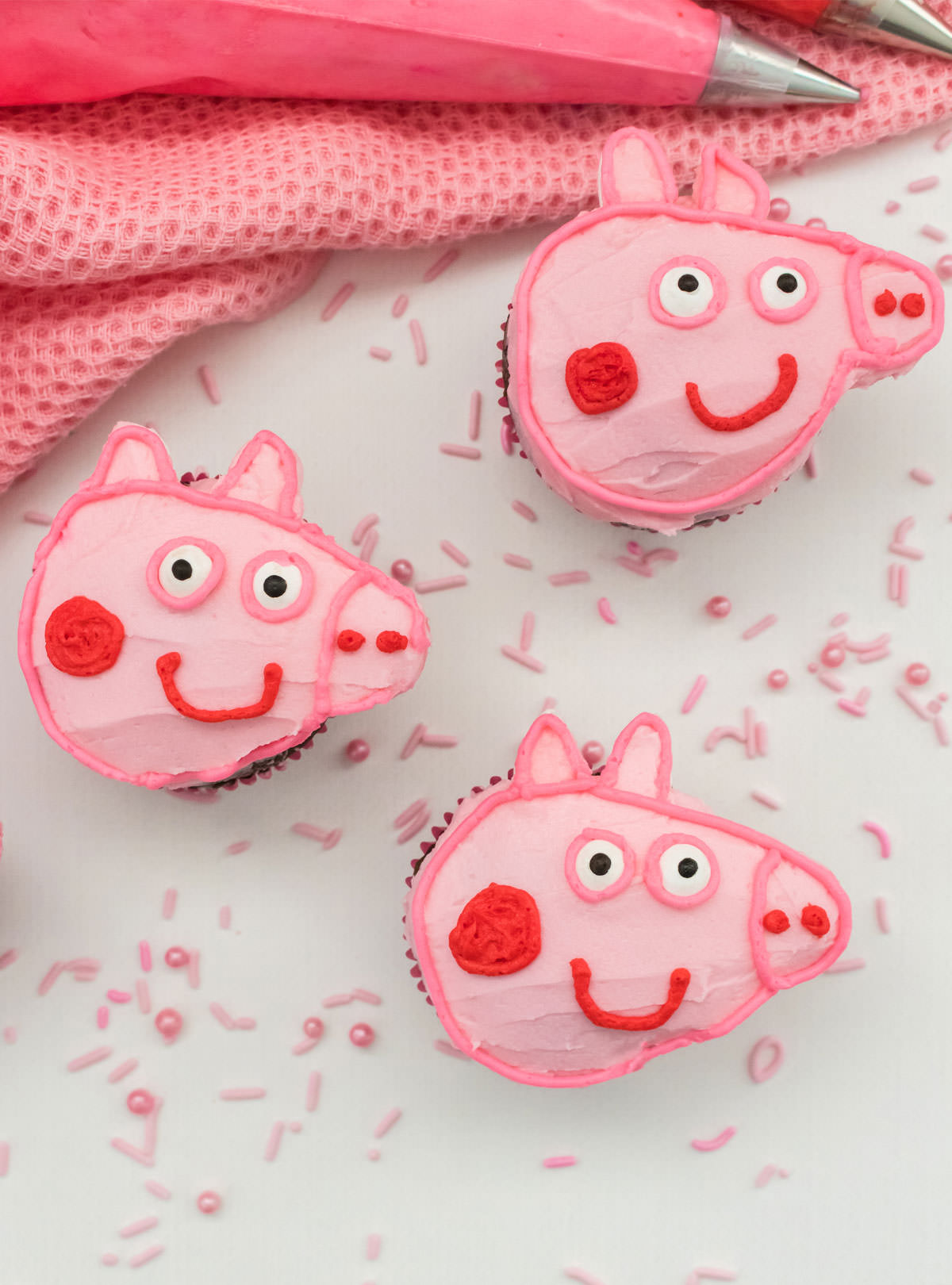 Closeup on three Peppa Pig Cupcakes sitting on a white surface, surrounded by pink sprinkles, in front of a pink table linen and pink frosting.