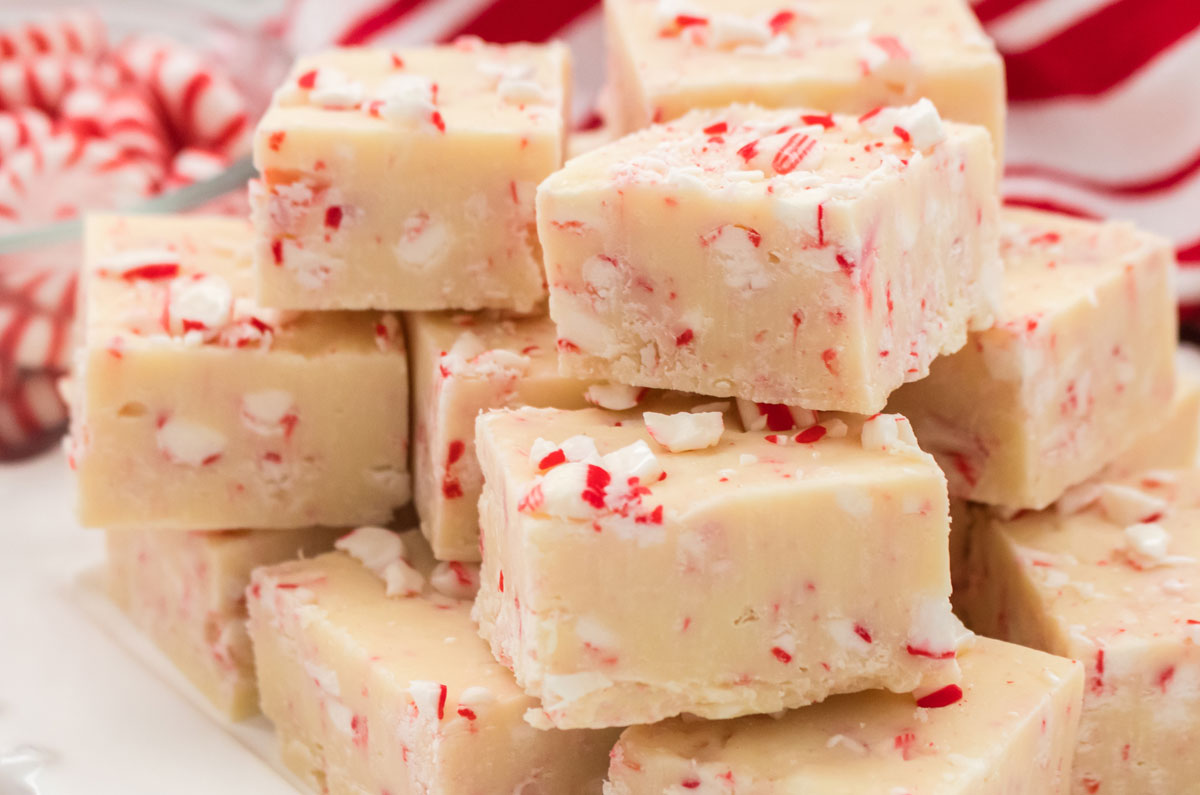 Closeup on a stack of Peppermint Fudge candies sitting on a white plate  with a glass bowl filled with Peppermint Candies in the background.