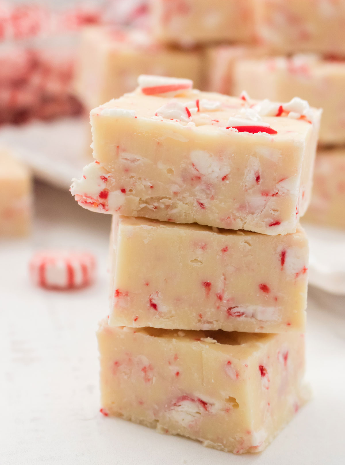 Closeup on a stack of three Peppermint Fudge candies sitting on a white table in front of a serving platter filled with Peppermint Fudge.
