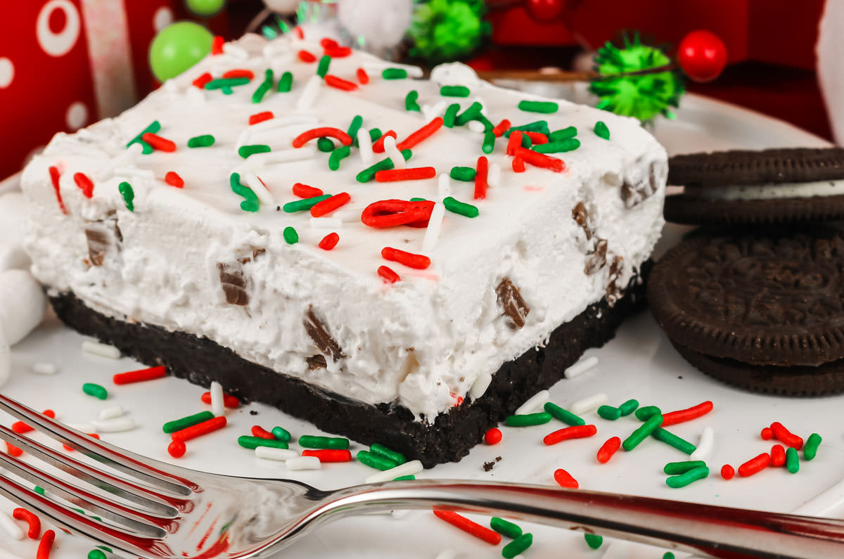 Closeup of a piece of Peppermint Marshmallow No-Bake Dessert sitting on a white plate surrounded by Oreo Cookies and Christmas sprinkles.