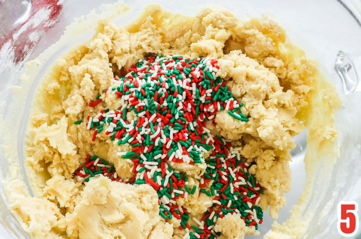 Closeup on a clear mixing bowl filled with sugar cookie dough and Christmas sprinkles.