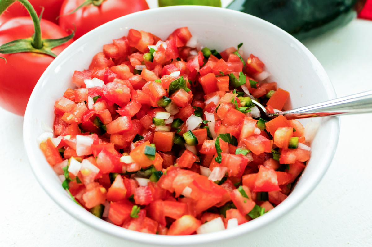 Closeup on a white bowl filled with Pico de Gallo sitting on a white table in front of tomatoes on the vine and a pablono pepper.
