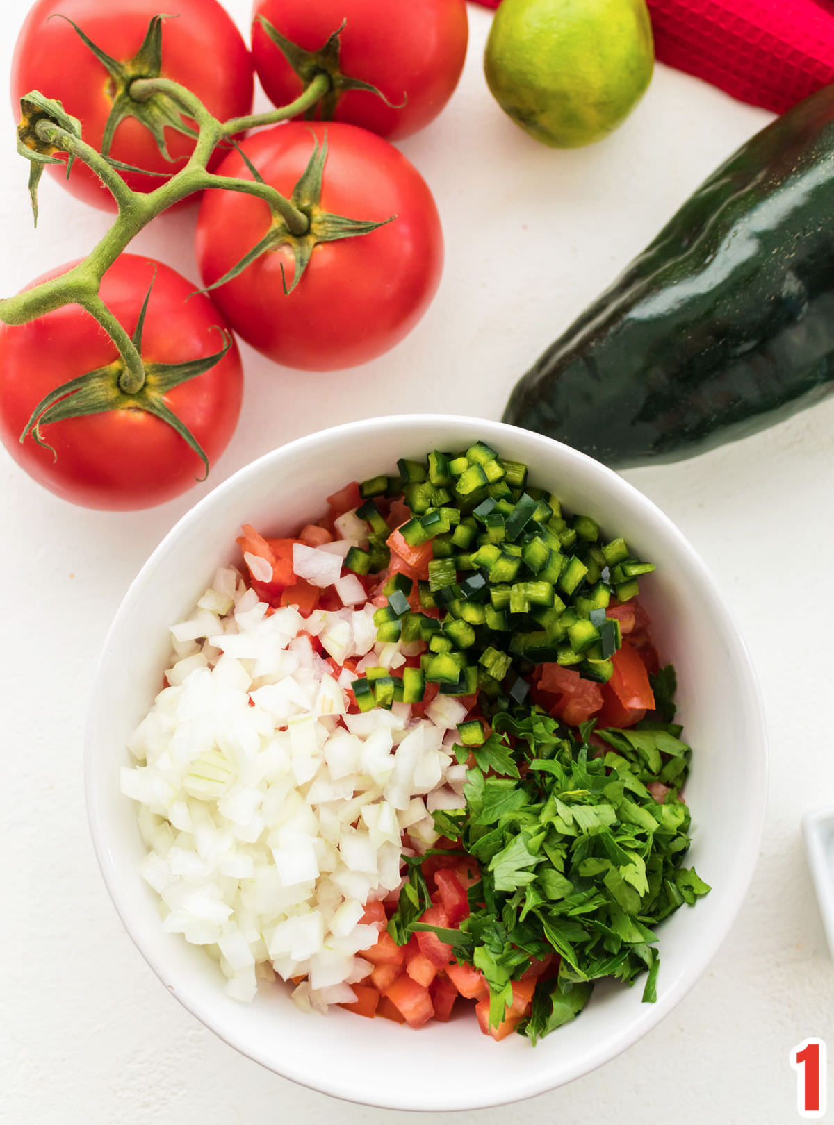 White bowl filled with diced tomatoes, onions, pablono peppers and cilantro sitting in front of tomatoes on the vine, lime and a pepper.