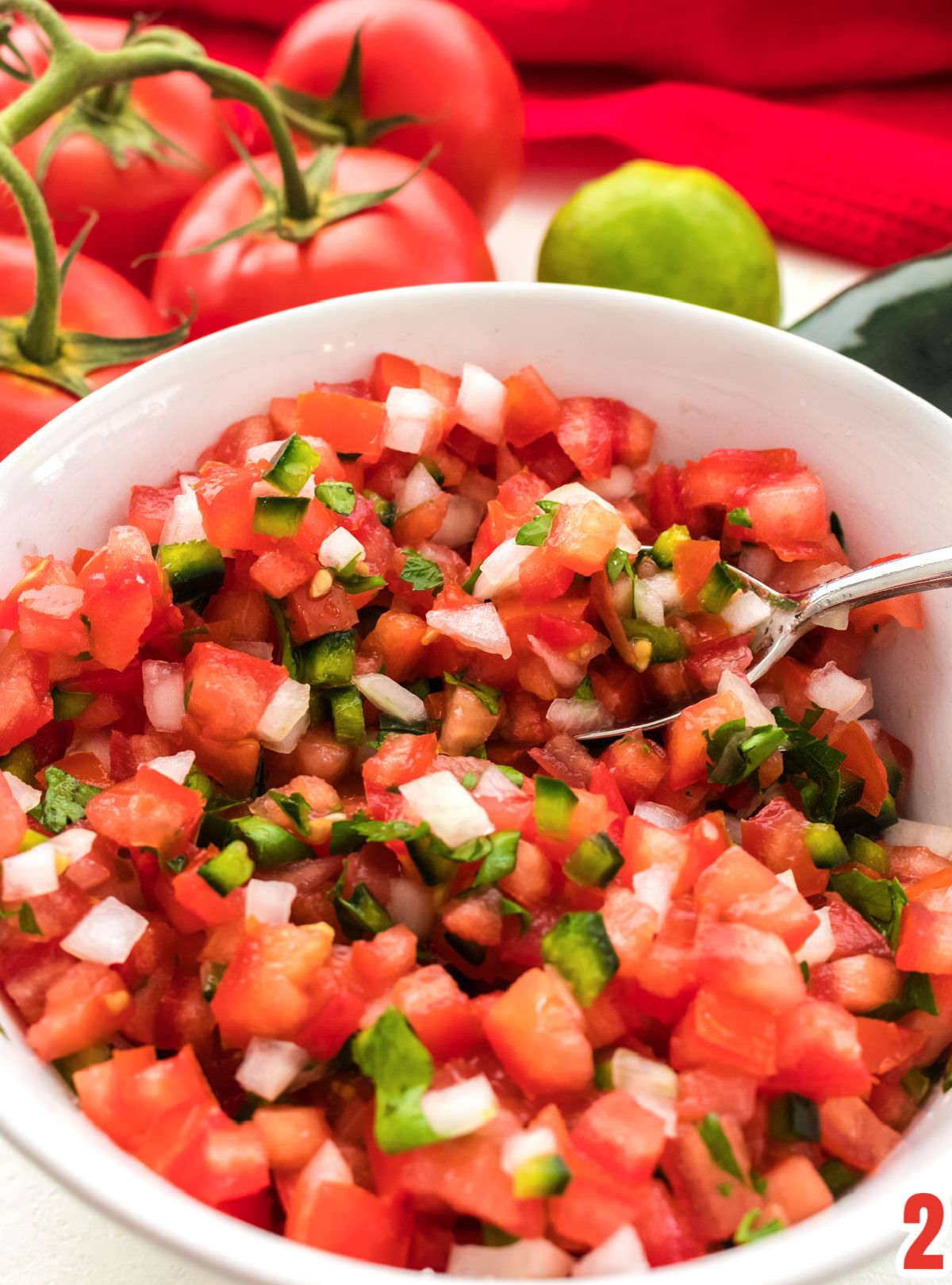 Closeup on a white serving bowl filled with Pico de Gallo with tomatoes, a lime and a pepper in the background.