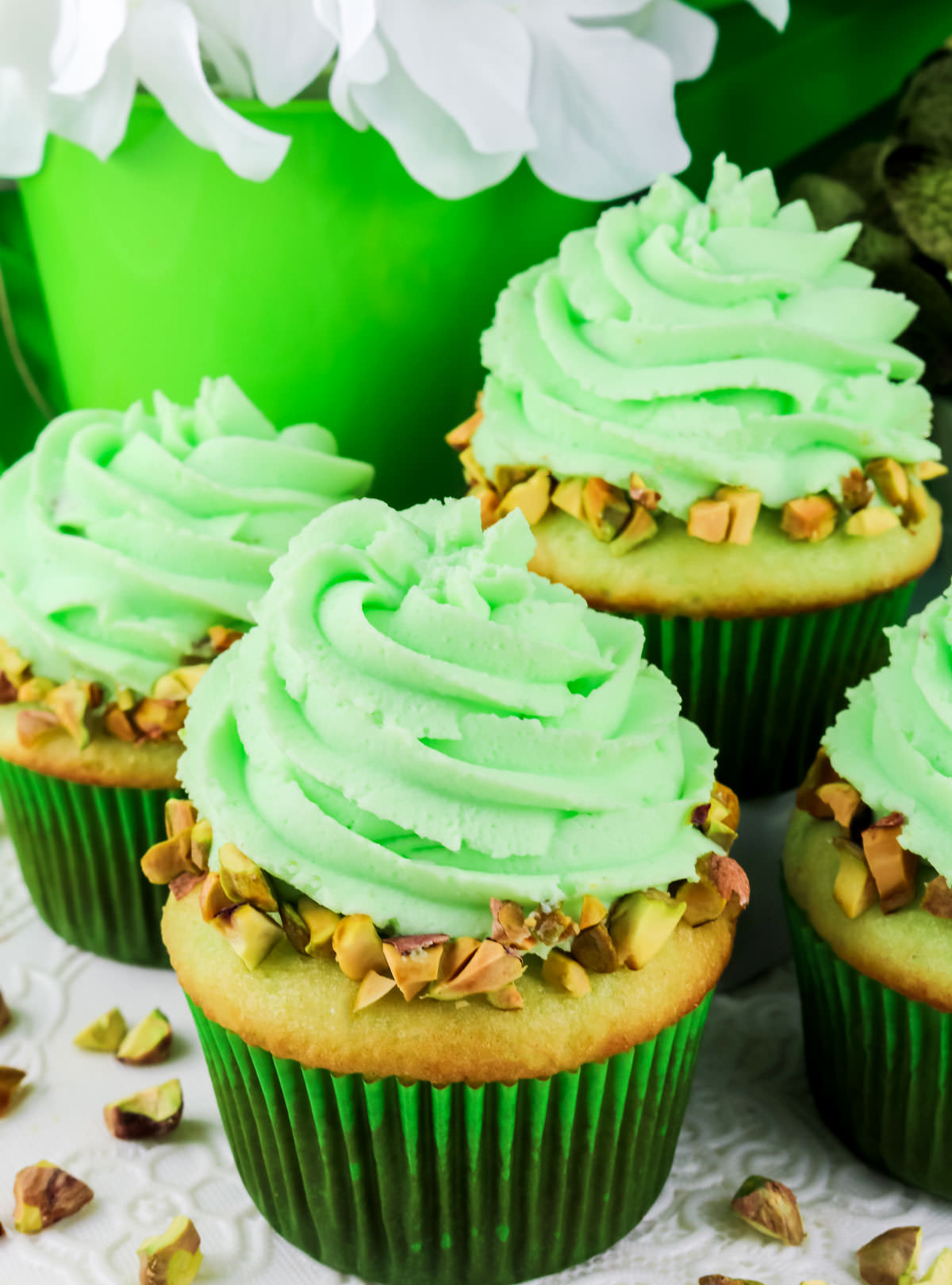 Four Pistachio Cupcakes with Pistachio Whipped Cream Frosting sitting on a white table surrounded by chopped Pistachio nuts.
