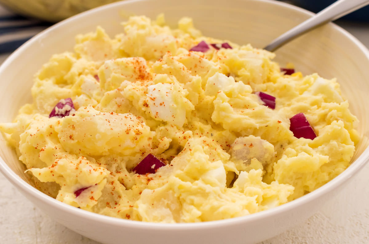 Close up on a white bowl filled with Classic Homemade Potato Salad sitting on a white table.