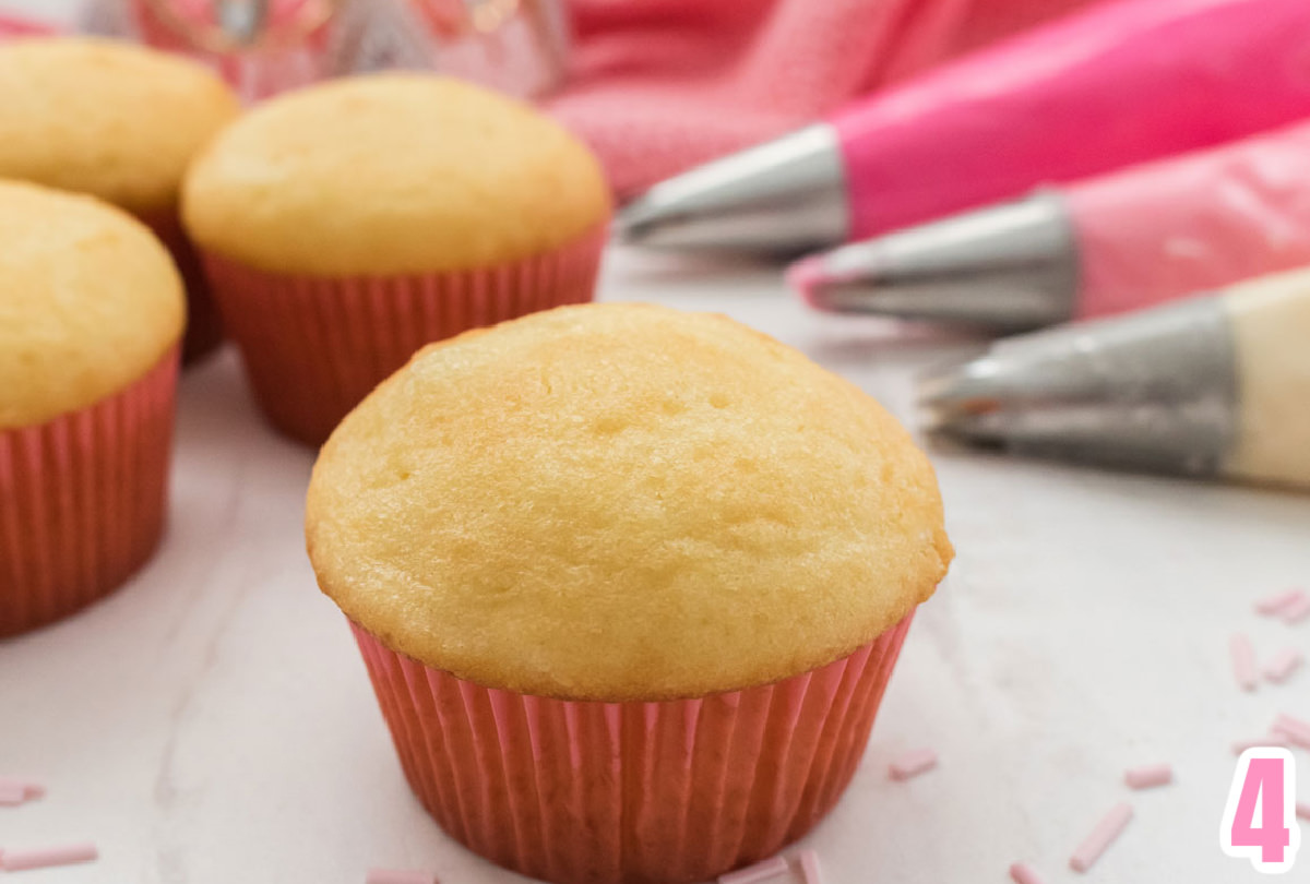 Closeup on a vanilla cupcake with a pink cupcake liner sitting on a white table next to more cupcakes and pink frosting in decorating bags.