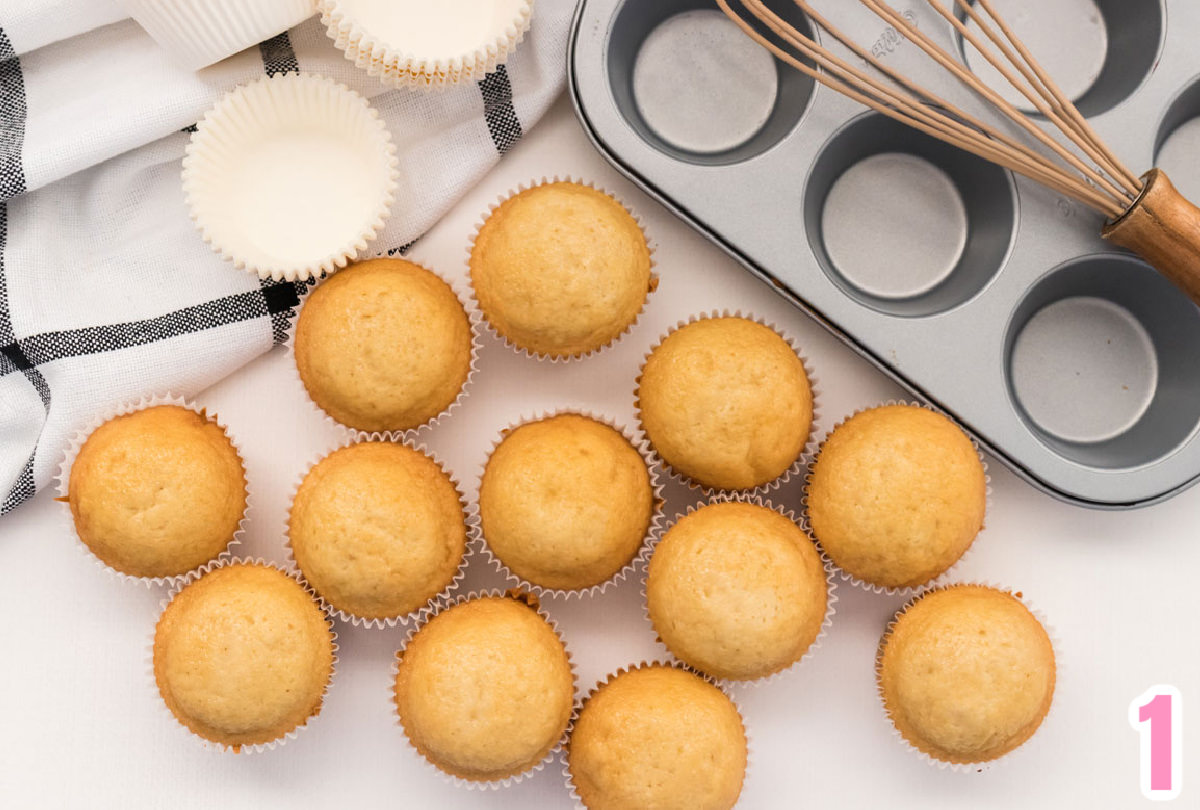 Closeup of a dozen vanilla cupcakes sitting on a white table surrounded by a cupcake tin and a black and white kitchen towel.