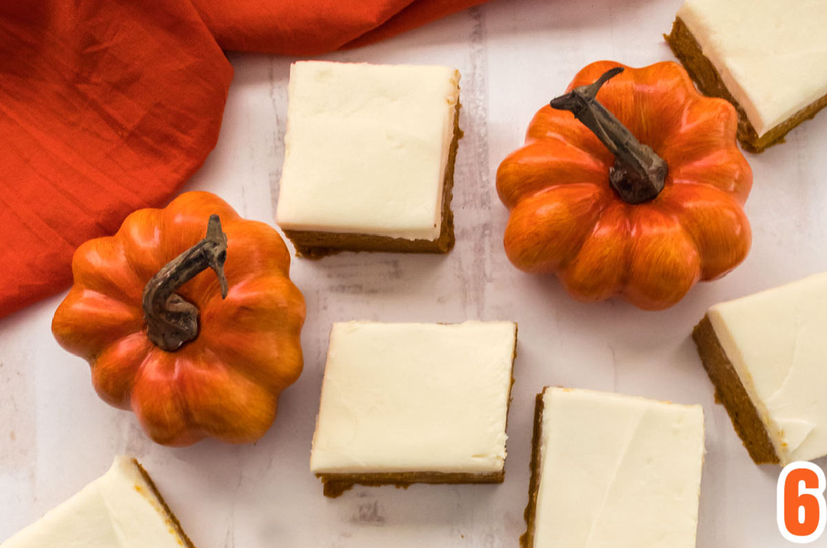 Overhead shot of five frosted Pumpkin Bars sitting on a white surface surrounded by mini pumpkins and an orange kitchen towel.
