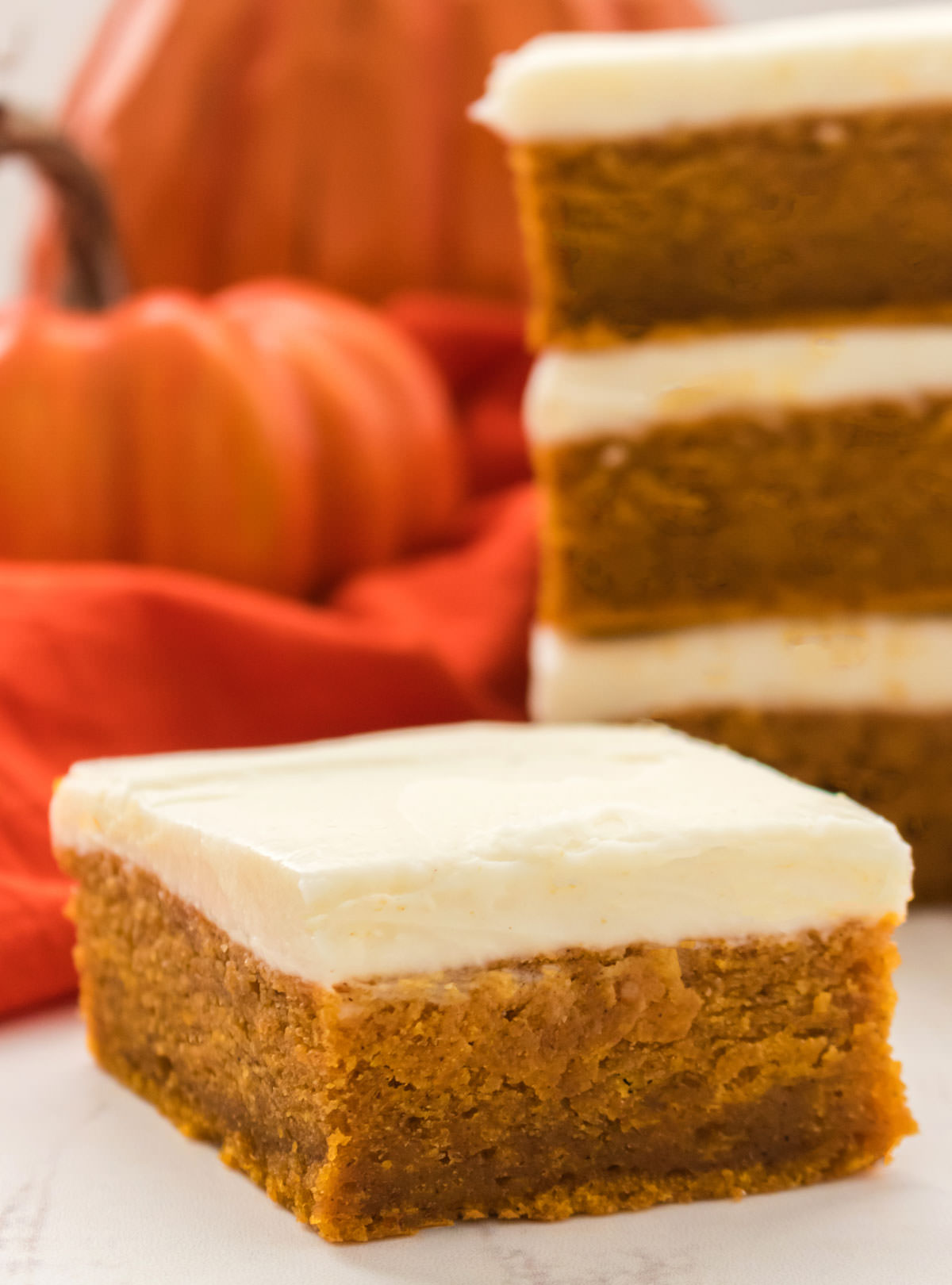 Closeup on a Pumpkin Bar with Cream Cheese Frosting sitting on a white surface in front of a stack of Pumpkin Bars and a Pumpkin decoration.