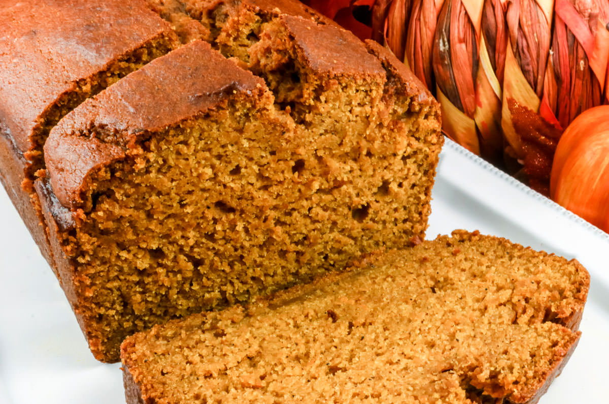 Closeup on a white serving platter filled with a loaf of Homemade Pumpkin Bread with a couple of pieces sliced and next to fall pumpkin decorations.