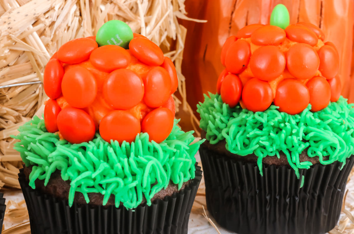 Closeup on two Pumpkin Patch Cupcakes sitting in front of a mini bale of hay and a pumpkin decoration.