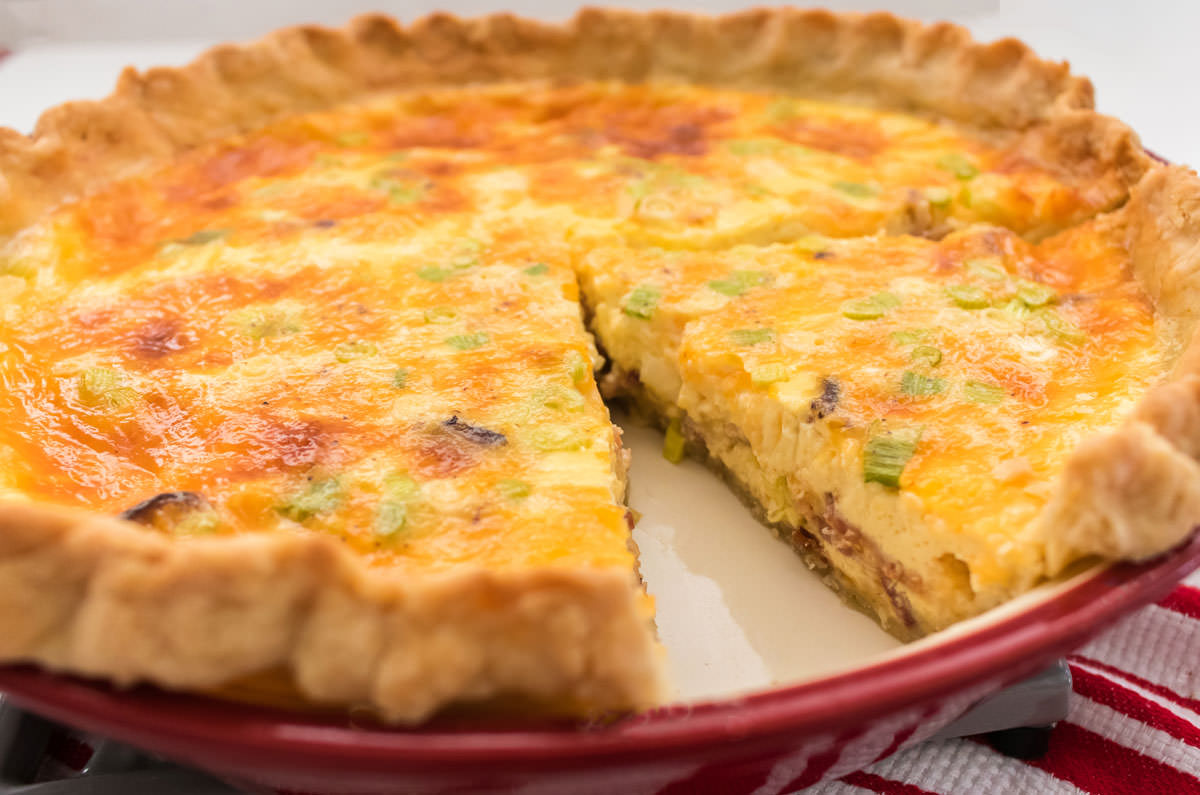 A closeup of a red pie plate of Quiche with one piece taken out.