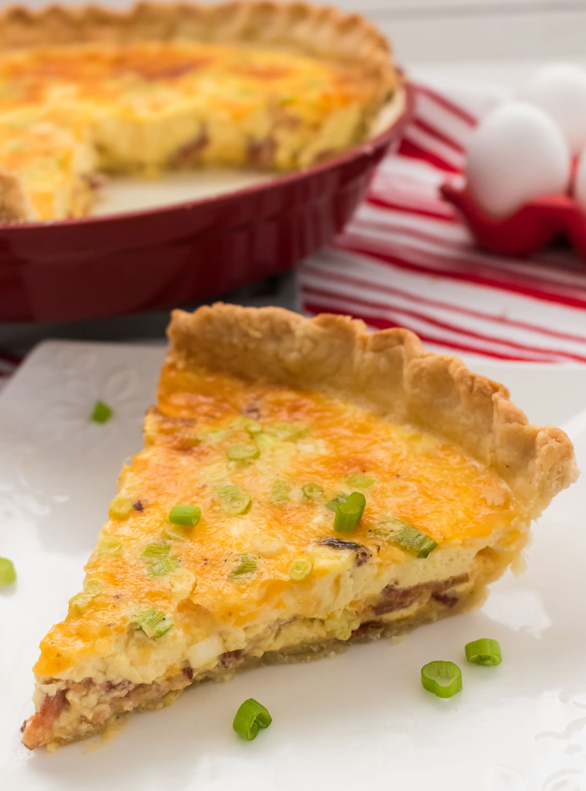 A closeup of a piece of Quiche Lorraine on a white plate sitting in front of the rest of the plate of quiche.